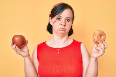 Down sendromlu esmer kadın elinde kırmızı elma ve çörek tutarken yüzünde ciddi bir ifadeyle rahatça oturuyor. Basit ve doğal bir şekilde kameraya bakıyorsun.. 