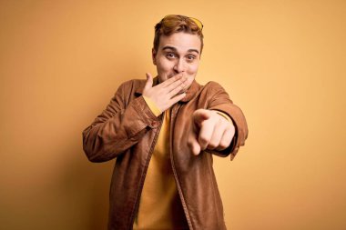 Sarı arka planda rahat deri ceket giyen genç, yakışıklı kızıl adam sana gülüyor, eli ağzında kamerayı işaret ediyor, utanç ifadesi takınıyor.