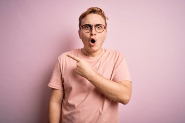 İzole edilmiş pembe arka planda dikilen sıradan tişörtlü, genç, yakışıklı, kızıl saçlı bir adam. Parmakla yüzünü göstermiş, ağzı açık, hayret dolu bir ifade..
