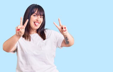Genç ve cüsseli bir kadın günlük kıyafetler giyip gülümsüyor ve iki elini de zafer işareti yaparken gösteriyor. İki numara.. 
