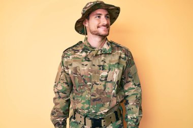 Kamuflaj üniforması giyen beyaz bir adam yüzünde gülümsemeyle, doğal bir ifadeyle yan yana bakıyor. Kendinden emin gülen. 