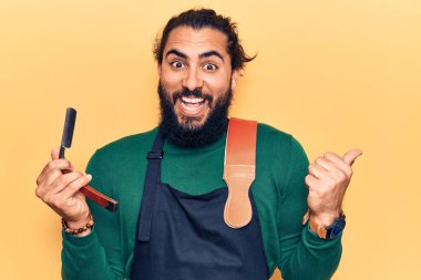 Önlük takan genç Arap adam berber jileti tutuyor baş parmağını yana doğru kaldırıyor ve ağzı açık gülümsüyor. 