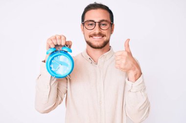 Çalar saati tutan yakışıklı adam mutlu ve pozitif gülümsüyor, başparmağıyla mükemmel ve onay işareti yapıyor. 