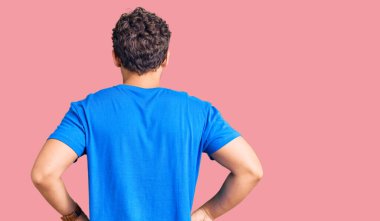 Kıvırcık saçlı, sıradan kıyafetler giyen, kolları vücudunda geriye doğru bakan genç, yakışıklı bir adam. 