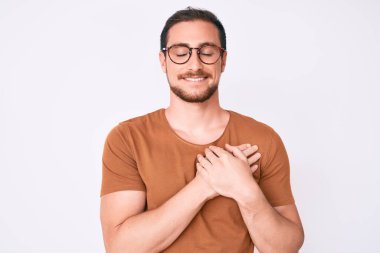 Gündelik kıyafetler ve gözlük takan genç yakışıklı adam elleri göğsünde, gözleri kapalı ve yüzünde minnet dolu bir jest ile gülümsüyor. sağlık kavramı. 