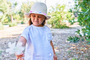 Yazlık şapkalı sevimli sarışın çocuk mutlu gülümsüyor. Yüzünde gülümsemeyle parkın etrafında çiçeklerle dikiliyordu.