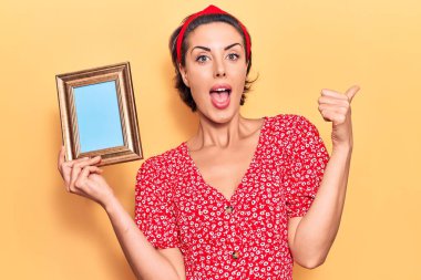 Elinde boş bir çerçeve tutan genç ve güzel bir kadın baş parmağını yana doğru uzatıyor ve ağzı açık gülümsüyor. 