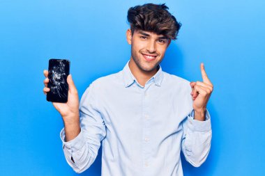 Kırık akıllı telefon tutan genç İspanyol adam kırık ekranı gösteriyor. Gülümsüyor, eli ve parmağı yandan işaret ediyor. 
