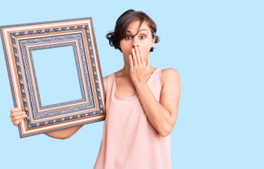 Kısa saçlı güzel bir kadın boş bir çerçeve ile ağzını eliyle kapatıyor, şokta ve hata yapmaktan korkuyor. şaşırmış ifade 