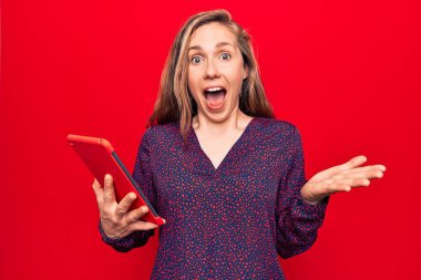 Güzel sarışın kadın, izole edilmiş kırmızı arka plan üzerinde tablet kullanıyor. Mutlu bir gülümsemeyle başarıyı kutluyor. Ve kazanan yüz ifadesi kaldırılıyor. 