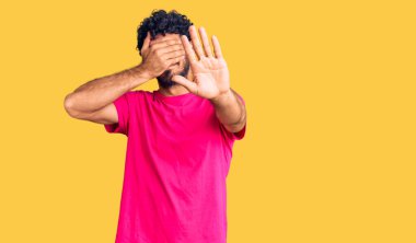 Kıvırcık saçlı yakışıklı genç adam ve sıradan pembe tişörtler giyen, ellerle gözlerini kapatan, üzgün ve korku ifadeli hareketler yapan ayı. utangaç ve olumsuz kavram. 