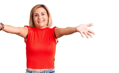 Sıradan kıyafetler giyen genç sarışın kadın kameraya bakıyor ve sarılmak için kollarını açmış gülümsüyor. Mutluluğu kucaklayan neşeli bir ifade.. 