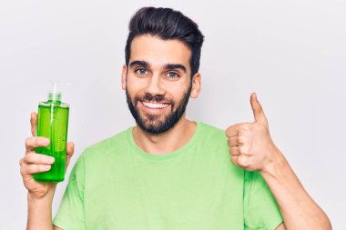 Sakallı genç yakışıklı adam aloe vera kremi şişesi giyiyor mutlu ve pozitif gülümsüyor, başparmağıyla mükemmel ve onay işareti yapıyor. 