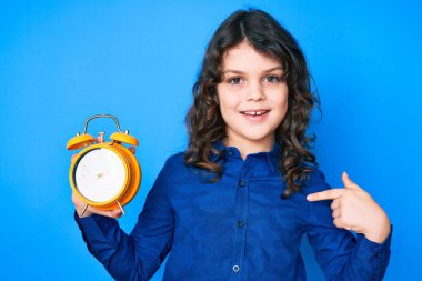 Uzun saçlı, sevimli İspanyol çocuk çalar saati tutuyor. Gülümseyen mutlu ve gururlu birini işaret ediyor. 