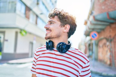 Genç beyaz adam mutlu bir şekilde gülümsüyor. Kulaklık takıyor ve şehirde yürüyor..