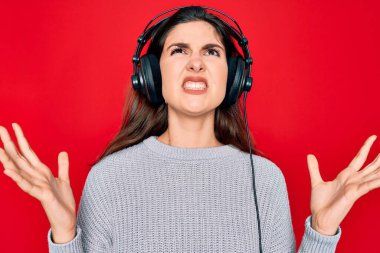 Modern kulaklık takan genç ve güzel bir kız kırmızı arka planda müzik dinliyor deli gibi bağırıyor ve agresif bir ifadeyle bağırıyor ve kollarını kaldırıyor. Hayal kırıklığı konsepti.