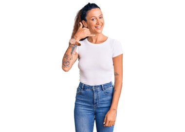 Dövmeli, beyaz tişörtlü genç bir İspanyol kadın. Gülümsüyor. El ve parmaklarıyla telefonla konuşuyor gibi. iletişim kavramları. 