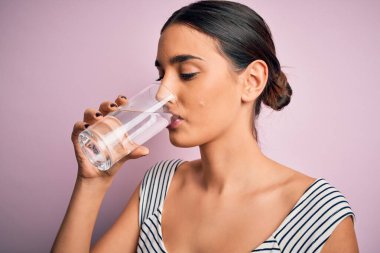 Genç, güzel esmer kadın, ferahlamak için bir bardak sağlıklı su içiyor. İzole edilmiş pembe arka planda duruyor.