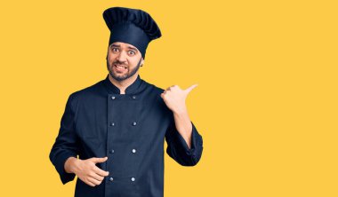 Pişirme üniforması giyen, gülümseyen, gülümseyen ve baş parmağıyla yan tarafı gösteren İspanyol genç adam.. 