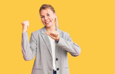 Zarif bir ceket giymiş genç, sarışın bir kadın. Arka tarafı işaret ediyor. Baş parmakları havada, kendine güvenen. 