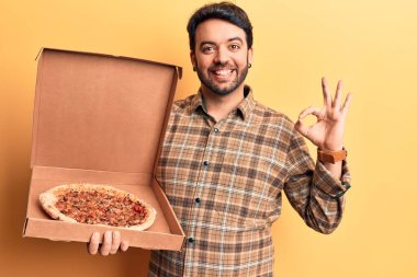 Elinde pizza kutusu tutan genç İspanyol adam parmaklarıyla imza atıyor, gülümseyerek mükemmel bir sembol oluşturuyor. 