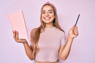 Genç, güzel, sarışın bir kadın elinde kitap ve kalemle mutlu ve havalı bir gülümsemeyle gülümsüyor. diş gösterme. 