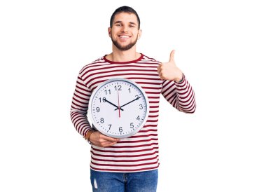 Büyük saati tutan yakışıklı genç adam mutlu ve pozitif gülümsüyor, başparmağıyla mükemmel ve onay işareti yapıyor. 