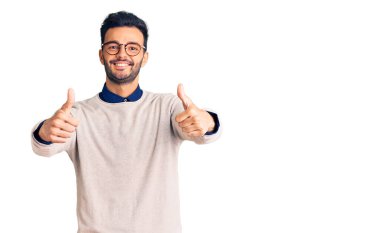 Zarif kıyafetler ve gözlük takan genç, yakışıklı İspanyol bir adam. Elle olumlu bir jest yapıyor. Gülümsüyor ve başarı için seviniyor. kazanan jest. 