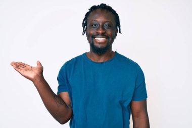 Saçları örgülü, neşeli gülümseyen, avuç içiyle kameraya bakan Afro-Amerikan genç adam.. 