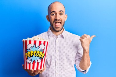 Genç ve kel bir adam elinde patlamış mısırla başparmağıyla yan tarafa bakıyor mutlu bir şekilde gülümsüyor. 