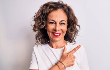 Orta yaşlı güzel esmer kadın beyaz arka planda dikilen sıradan tişörtler giyiyor. Elleri ve parmaklarıyla neşeli bir şekilde işaret ediyor.