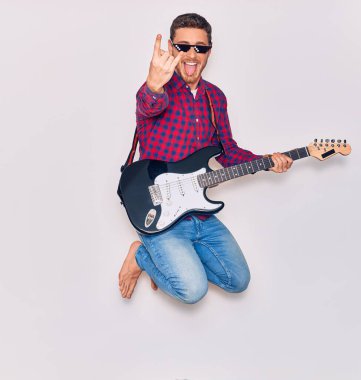 Serseri hayatı takan genç İspanyol sanatçı ağzı ve dili açık güneş gözlüğü takıyor. Yüzünde tebessümle atlamak elektro gitar çalmak izole beyaz arka planda boynuz işareti yapmak.