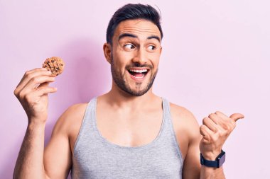 Sakallı, kolsuz tişörtlü, çikolatalı kurabiye yiyen, başparmağını kaldırıp gülümseyen, ağzı açık genç yakışıklı bir adam.