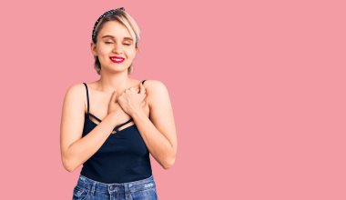 Genç, güzel, sarışın bir kadın sıradan kıyafetler giyiyor. Elleri göğsünde, gözleri kapalı gülümsüyor ve yüzünde minnet ifadesi var. sağlık kavramı. 