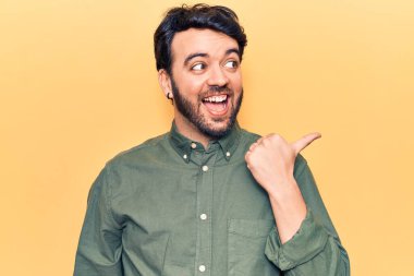 Gündelik kıyafetler giyen, gülümseyen, gülümseyen ve başparmağıyla yan tarafı gösteren İspanyol genç adam.. 