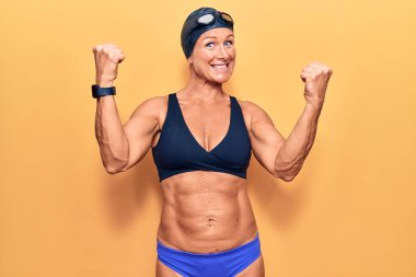 Orta yaşlı sarışın kadın bikini giyiyor, yüzme şapkası takıyor ve Google 'da gururla bağırıyor zaferi ve başarıyı kollarını kaldırarak kutluyor. 