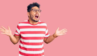 Yakışıklı Latin Amerikalı genç adam gündelik kıyafetler ve gözlük takıyor deli gibi bağırıyor ve bağırıyor agresif bir ifade ve kollar havada. hüsran kavramı. 