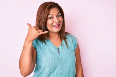 Orta yaşlı esmer, sıradan kıyafetler giyen, gülümseyen, el ve parmaklarıyla telefon hareketi yapan telefonda konuşan Latin bir kadın. iletişim kavramları. 