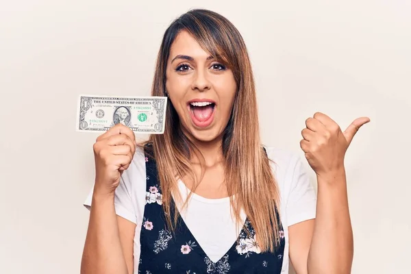 Joven Hermosa Mujer Sosteniendo Dólar Señalando Pulgar Hacia Lado Sonriendo —  Fotos de Stock