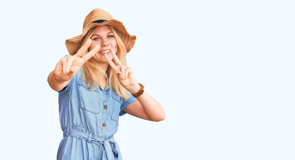 若い美しいブロンドの女性は夏の帽子をかぶって 指が勝利のサインをしているのを示すカメラを見て笑ってドレスします — ストック写真