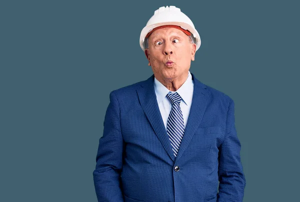 Senior handsome grey-haired man wearing suit and architect hardhat making fish face with lips, crazy and comical gesture. funny expression.