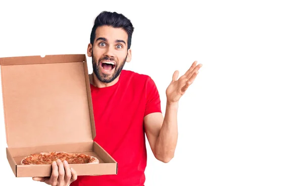 年轻英俊的男人留着胡子 拿着纸板箱和意大利披萨庆祝胜利 脸上带着快乐的笑容和高傲的双手 — 图库照片