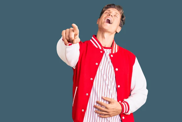 Young handsome man wearing baseball uniform laughing at you, pointing finger to the camera with hand over body, shame expression