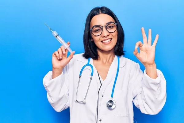 Young beautiful latin woman wearing doctor stethoscope holding syringe doing ok sign with fingers, smiling friendly gesturing excellent symbol