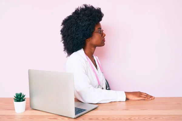 年轻的非洲裔美国女人戴着医生听诊器 一边用电脑笔记本电脑工作一边看 带着自信的微笑放松自己的姿态 — 图库照片