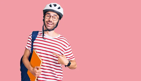 Young hispanic man wearing student backpack and bike helmet holding binder smiling happy pointing with hand and finger