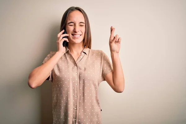 白い背景のジェスチャーの指でスマートフォンで会話をしている若い美しい女性が希望と目を閉じて笑顔を交わした 幸運と迷信の概念 — ストック写真