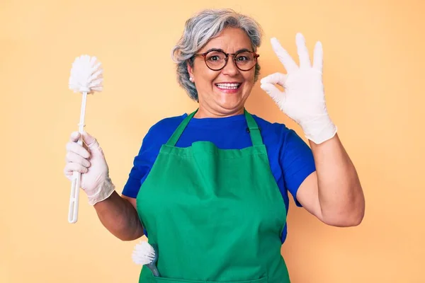 Senior hispanic woman wearing apron holding brush doing ok sign with fingers, smiling friendly gesturing excellent symbol