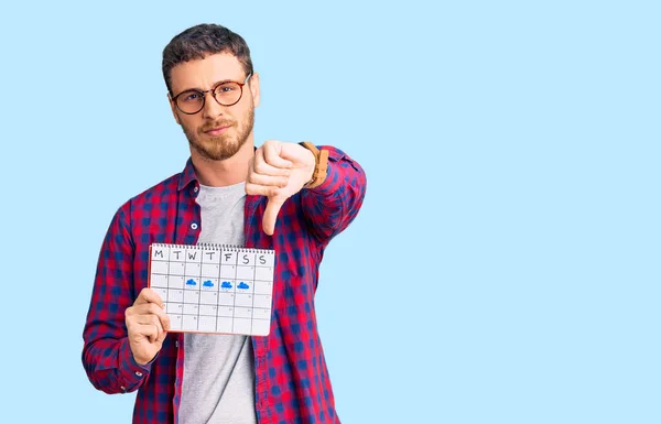 Handsome young man with bear holding travel calendar with angry face, negative sign showing dislike with thumbs down, rejection concept