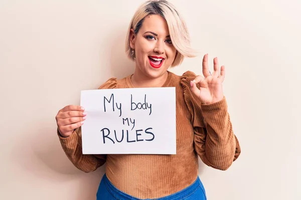 Blonde plus size woman asking for women rights holding paper with my body my rules message doing ok sign with fingers, smiling friendly gesturing excellent symbol